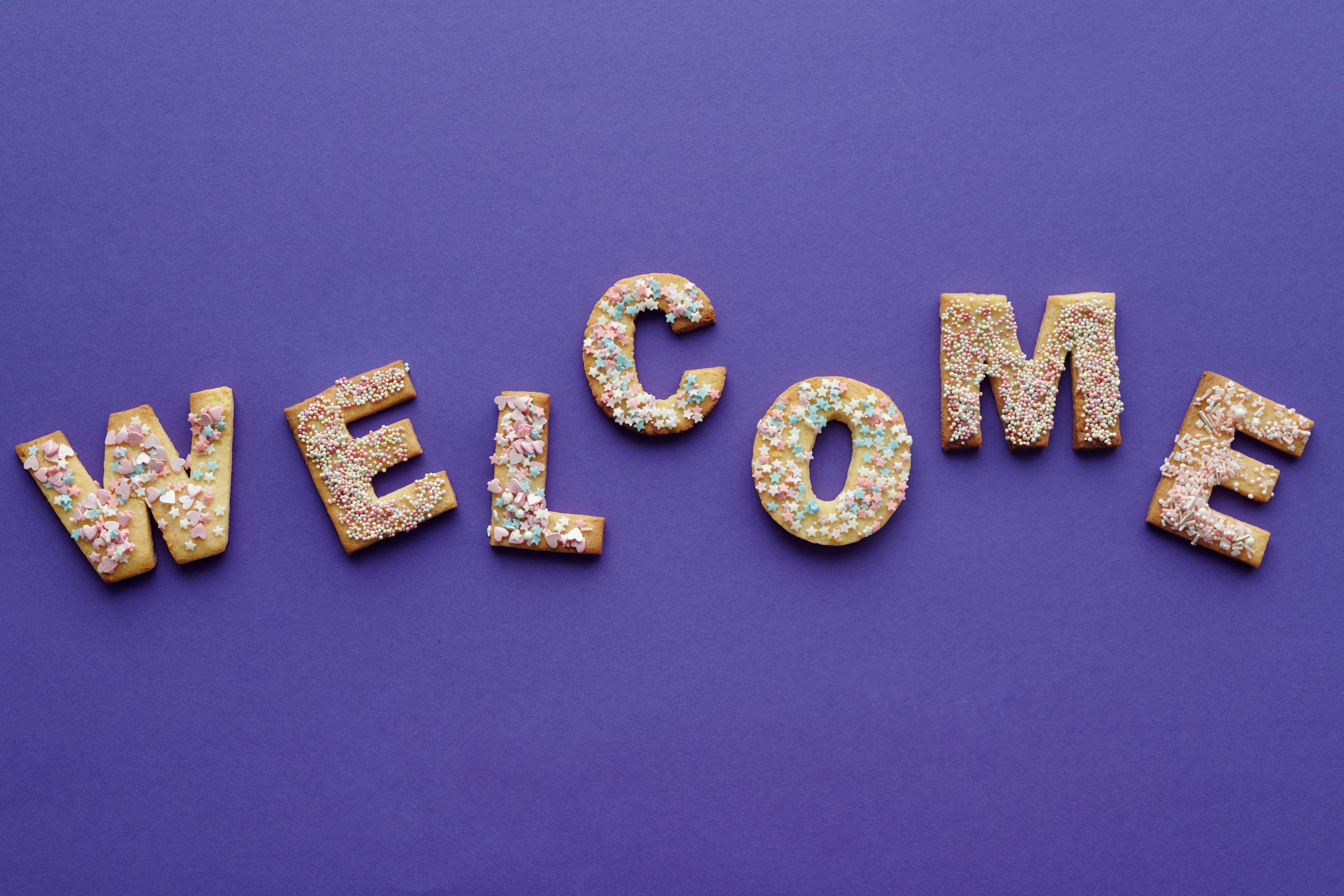 Colorful sprinkled cookies spelling 'welcome' on purple backdrop, perfect for celebrations.