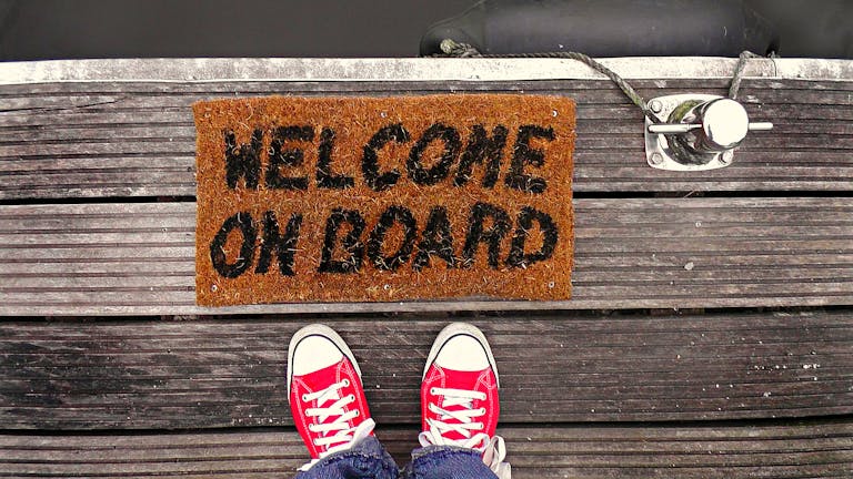 A welcome mat on a wooden dock with a pair of red sneakers visible from above.